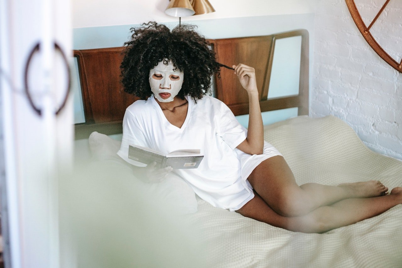 Young black lady reading book on bed during skin care routine at home.